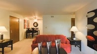 This single-floor space features an open-concept design that seamlessly connects the living and dining areas, creating a cohesive and functional layout. The living area is anchored by plush, comfortable seating and is complemented by warm, neutral-toned carpeting underfoot. A ceiling fan with an integrated light fixture provides both airflow and ambient lighting overhead, contributing to the room's practical design.

The dining area, visible from the living space, accommodates a full dining set with multiple chairs, making it well-suited for everyday meals or casual gatherings. Wall-mounted décor adds a pop of color and personality to the space. Transition areas include doorways that lead to additional rooms, suggesting a practical floor plan throughout. Ventilation is well-considered, with ceiling vents distributed across the space. The warm lighting from a table lamp in the corner adds to the inviting ambiance. Overall, the space presents a comfortable and efficiently arranged interior with a cohesive warm color palette throughout the main living areas.