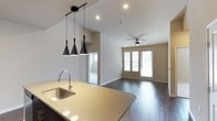 This well-appointed single-floor space showcases a thoughtfully designed open-concept layout that seamlessly connects the kitchen and living areas. The kitchen features a large quartz countertop island with an undermount sink, offering ample workspace and a natural gathering point. Three striking matte black cone-shaped pendant lights hang above the island, adding a modern, sophisticated touch to the space.

The living area benefits from vaulted ceilings that enhance the sense of openness and airiness throughout. A ceiling fan is centrally positioned to ensure comfortable air circulation. The space is bathed in natural light, courtesy of a set of glass French doors flanked by windows at the far end of the living area, offering a bright and welcoming atmosphere.

Rich dark hardwood flooring extends throughout, providing a warm contrast to the neutral, light-toned walls and ceilings. Recessed lighting complements the pendant fixtures, ensuring the space is well-lit at all times. The clean, contemporary design is defined by its minimalist aesthetic, open flow, and high-quality finishes — making this a versatile and inviting space suitable for both relaxing and entertaining.