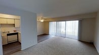 This single-floor space presents an open layout that seamlessly connects two distinct living zones, each with its own flooring material. The front area is finished with plush, neutral-toned carpeting, providing a comfortable and warm surface underfoot. Toward the back of the space, the flooring transitions to dark hardwood-style planks, adding a contrasting, more formal finish to the area.

A large sliding glass door dominates one wall, fitted with full-length vertical blinds that allow for flexible light control. The door allows an abundance of natural light to fill the interior, brightening the otherwise neutral-toned walls. A ceiling fan with an integrated light fixture is centrally positioned in the rear section, offering both air circulation and ambient lighting.

The walls are finished in a clean, light neutral tone throughout, contributing to an open and airy feel. White baseboards frame the perimeter of the space neatly. The overall layout is functional and straightforward, offering a blank canvas well-suited for a variety of furniture arrangements and interior styles.
