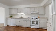 This single-floor home offers a clean, functional layout designed for comfortable everyday living. The interior is defined by a consistent design palette of crisp white walls and rich, dark hardwood-style flooring that runs through the main living and kitchen areas, creating a warm contrast throughout.

The kitchen is well-equipped with ample light wood cabinetry providing generous storage above and below the counter, a double sink, a full suite of white appliances including a dishwasher and electric range, and a built-in range hood. The kitchen opens toward the living area, enhancing the sense of connectivity between spaces.

The living and bedroom areas feature plush wall-to-wall carpeting, lending a comfortable, quiet feel underfoot. Bedrooms are naturally lit by sliding windows that frame views of lush greenery outside. A spacious walk-in closet with built-in shelving and a hanging rod offers excellent storage for the primary bedroom.

The bathroom includes a full soaking tub with white tile surround, mosaic tile flooring, and a bright vanity with a large mirror. A dedicated laundry room with a full-size washer and dryer adds practical convenience. The central hallway connects all areas of the home efficiently, giving the overall layout a cohesive, easy-to-navigate flow.