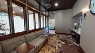 Located at 8585 La Mesa Blvd in La Mesa, San Diego, this single-floor professional space combines classic architectural character with modern office functionality. The interior is defined by a striking nature-inspired patterned carpet in warm earth tones of brown, gold, and cream, which flows throughout the space and anchors the overall design aesthetic.

A prominent lounge area features a comfortable gray sectional sofa paired with a dark wood side table, positioned alongside a dramatic wall of tall, dark wood-framed windows. These windows flood the space with abundant natural light while offering views of the surrounding neighborhood. The elevated ceiling height in this area, accented by a dark wood paneled ceiling, lends a sense of grandeur and distinction.

The layout includes multiple connected zones, with a secondary seating alcove featuring an arched window and additional lounge furniture, ideal for informal meetings or waiting areas. A dedicated workstation area with a desk and ergonomic seating supports day-to-day office functions. Double glass-paneled interior doors with a classic design provide access between areas, enhancing both connectivity and visual openness throughout the space. The neutral gray wall palette complements the rich wood trim and warm flooring, creating a professional yet inviting atmosphere suited for client-facing business operations.
