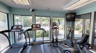 This fitness center is a single-floor facility designed to support a well-rounded workout routine. The space is organized into distinct functional zones, beginning with a dedicated cardio area housing multiple treadmills and elliptical trainers arranged along a wall of large windows. These windows flood the area with natural light and offer views of the surrounding greenery and outdoor spaces, creating an open, energizing atmosphere during workouts. A wall-mounted television is positioned nearby for added convenience during cardio sessions.

The strength training area extends further into the facility and features a Body-Solid cable machine capable of supporting a variety of resistance exercises, along with a weight bench for free weight and strength routines. Large glass panel walls partition sections of the space, maintaining visual openness throughout. The flooring transitions between carpet tile and rubberized mat areas, providing appropriate surfaces for different exercise types.

Overhead, recessed panel lighting and drop-ceiling fixtures ensure consistent illumination throughout the facility. Security cameras are installed for safety monitoring. An exterior door with a white railing deck provides direct access to the outdoors. The walls are painted in a soft teal tone, contributing to a clean and motivating environment.