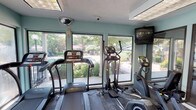 This fitness center is a single-floor facility designed to support a well-rounded workout routine. The space is organized into distinct functional zones, beginning with a dedicated cardio area housing multiple treadmills and elliptical trainers arranged along a wall of large windows. These windows flood the area with natural light and offer views of the surrounding greenery and outdoor spaces, creating an open, energizing atmosphere during workouts. A wall-mounted television is positioned nearby for added convenience during cardio sessions.

The strength training area extends further into the facility and features a Body-Solid cable machine capable of supporting a variety of resistance exercises, along with a weight bench for free weight and strength routines. Large glass panel walls partition sections of the space, maintaining visual openness throughout. The flooring transitions between carpet tile and rubberized mat areas, providing appropriate surfaces for different exercise types.

Overhead, recessed panel lighting and drop-ceiling fixtures ensure consistent illumination throughout the facility. Security cameras are installed for safety monitoring. An exterior door with a white railing deck provides direct access to the outdoors. The walls are painted in a soft teal tone, contributing to a clean and motivating environment.