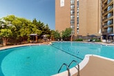 Located at 250 S Whiting St in Alexandria, Virginia, this outdoor amenity area serves as a central gathering space for the residential community. The centerpiece is a generously sized freeform swimming pool with a distinctively curved, multi-lobed shape, finished with crisp white coping and clear turquoise water. Lane markers stretch across the pool, and built-in entry steps with stainless steel handrails provide easy access. The expansive pool deck is finished in a warm tan concrete surface, offering ample room for lounging and circulation. Sunbathing areas are equipped with reclining lounge chairs, and shaded retreats are provided by multiple large market-style umbrellas and a fabric canopy cabana. A lifeguard stand is positioned poolside, underscoring the well-maintained nature of the facility. An adjacent patio area features outdoor dining tables and chairs beneath additional shade umbrellas, ideal for casual gatherings. Ornamental iron fencing encloses the space, lending a sense of privacy and security. Mature trees and landscaped greenery line the perimeter, creating a lush, park-like backdrop. The area sits adjacent to the high-rise residential tower, with balconies overlooking the pool and open sky above, making it a bright and inviting outdoor retreat throughout the warmer months.