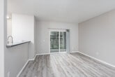 This single-floor residence offers a thoughtfully laid-out interior finished with a neutral, modern palette throughout. The open living area is anchored by light wood-look flooring and features a ceiling fan overhead, while a sliding glass door fills the space with natural light and provides direct access to an outdoor patio, offering views of surrounding greenery.

The kitchen is positioned adjacent to the living area and is equipped with stainless steel appliances, including a refrigerator, electric range, and over-the-range microwave. Light wood cabinetry pairs with speckled blue-gray countertops, offering functional workspace and ample storage.

The bedroom is a calm, comfortable retreat finished with plush wall-to-wall carpeting, a baseboard heater, and a window fitted with blinds that frame outdoor views. The bathroom features a tub-shower combination with tiled surround, a curved shower curtain rod, and light wood-look flooring that complements the rest of the unit. A second bathroom area includes a vanity with a gray countertop and additional storage. Throughout, the space maintains a clean, understated aesthetic with consistent neutral tones and practical finishes.