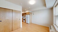 This efficient single-floor unit is thoughtfully laid out to maximize every square foot, comprising a living room, kitchen, bathroom, and two closets. Light hardwood flooring runs throughout, lending warmth and visual continuity across all spaces.

The living room is bright and open, anchored by a pair of large windows fitted with blinds that frame views of the surrounding neighborhood. A wall-mounted HVAC unit provides climate control, and the clean white walls create a neutral, welcoming ambiance.

The kitchen is well-appointed with warm honey-toned wood cabinetry extending from floor to ceiling, offering generous storage. A stainless steel sink, white electric range with coil burners, and a full-size refrigerator complete the functional setup, all set against crisp white countertops.

The bathroom is clean and simply finished, featuring a wood-toned vanity with an integrated sink, a wall mirror, and a combined tub and shower. Consistent light wood flooring ties the space to the rest of the unit.

Two closets provide practical storage solutions, including one with a distinctive natural wood-panel door. Recessed and flush-mount ceiling lighting throughout keeps the unit well-illuminated. The overall design is clean and functional, with a consistent palette of light wood and white tones.