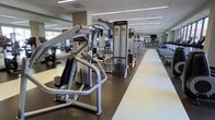 This fitness center offers a comprehensive and professionally designed workout environment spread across a single open floor. The space is outfitted with an extensive lineup of Life Fitness strength training machines, including cable systems, weight machines, and resistance equipment, all arranged in an organized and accessible layout. A dedicated rubberized flooring zone houses the strength equipment, providing both safety and stability during workouts, while a light-colored pathway runs alongside the equipment for easy navigation throughout the facility. The space benefits from a drop ceiling with recessed and linear fluorescent lighting, ensuring bright, even illumination throughout. Large windows along one side of the facility allow natural light to filter in, contributing to an energizing and open atmosphere. Mirrors line portions of the walls, enhancing the sense of space while allowing users to monitor their form. Rows of cardio machines are visible toward the far end of the floor, rounding out the facility's full-body training offerings. The overall design is clean, modern, and functional, reflecting a professional-grade fitness environment well-suited for a wide range of users and fitness goals.