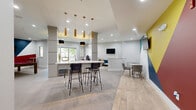 This stylish single-floor community space offers a well-appointed and energetic environment designed for socializing and recreation. The open-plan layout seamlessly connects the kitchen and living areas, creating a cohesive and inviting atmosphere throughout. Light-toned tile flooring in a herringbone pattern runs across the main gathering areas, adding a refined yet contemporary touch.

The kitchen zone features a large white countertop island with bar-height seating, a built-in beverage cooler, and a window that draws in natural light. A striking cluster of gold and black cylindrical pendant lights hangs above the island, serving as a bold focal point. Structural columns punctuate the space without disrupting the open flow.

The living area includes a tufted teal sofa accented with gold throw pillows, marble-topped coffee tables with gold legs, and a wall-mounted television — creating a comfortable lounge setting. Intimate seating nooks with round marble tables and red velvet chairs provide additional gathering spots throughout the space.

A dedicated game room features a full-size pool table with a red felt surface, wall-mounted cue storage, and high-top bistro tables. The standout design element across the space is a series of bold geometric accent walls in yellow, teal, navy, and red — lending a dynamic, contemporary energy to the overall aesthetic.