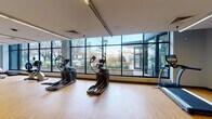 This fitness center offers a comprehensive workout environment designed to accommodate a wide range of exercise needs. The space is characterized by its open, elongated layout with light hardwood flooring throughout, creating a clean and energizing atmosphere. Large, floor-to-ceiling windows line the perimeter, flooding the interior with abundant natural light and providing open views of the surrounding streetscape and greenery, enhancing the overall workout experience.

The cardio zone is well-stocked with multiple elliptical trainers, treadmills, and a recumbent bike, all positioned along the windowed walls to take full advantage of the natural light and outdoor views. A multi-station cable and strength machine anchors the strength training area, accompanied by free weights, dumbbells, stability balls, and a flat exercise bench, offering versatility for resistance training routines.

Linear LED lighting runs along the ceiling, supplementing natural light and ensuring even illumination throughout. Structural columns punctuate the space without disrupting the open flow, while the neutral white and grey walls contribute to a clean, professional aesthetic. A water cooler and additional accessories are conveniently accessible within the space. The fitness center presents a well-rounded, functional facility suited for daily training.
