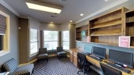 This single-floor business center is thoughtfully designed to balance productivity with comfort. The room features a built-in wooden workstation along one wall, equipped with dual computer monitors and keyboards, making it ideal for guests or members needing a dedicated workspace. Above the desk, open wood shelving provides convenient storage and display space, adding a warm, refined character to the room.

A charming bay window area anchors the opposite corner, fitted with elegant Roman shades and horizontal blinds that allow for adjustable natural light. Two patterned accent chairs are positioned beneath the windows, creating an inviting lounge nook perfect for reading or informal meetings. The greenery visible through the windows adds a pleasant outdoor connection to the interior.

The flooring features a bold geometric-patterned carpet in deep tones, which contrasts beautifully with the neutral gray walls and warm wood tones of the cabinetry. Crown molding and recessed lighting lend a polished, professional finish to the ceiling, while a central skylight panel enhances the overall brightness of the space. The overall ambiance is one of quiet efficiency and approachable elegance, well-suited for focused work in a welcoming environment.