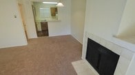 This single-story space offers a straightforward, functional layout with clearly defined living and kitchen areas. The living room is finished with wall-to-wall carpet in a neutral tan tone, creating a comfortable and understated foundation. A standout feature of this room is the fireplace positioned along one wall, framed by white tile surround and hearth, adding a classic focal point to the space.

The living area opens directly toward the kitchen, which is set slightly apart with hard flooring that contrasts the carpeted living space. The kitchen features wood-toned upper and lower cabinetry, a double sink positioned beneath a window that allows natural light into the space, and a refrigerator. A peninsula counter separates the kitchen from the living area, offering additional workspace and a natural transition between the two zones. A pendant light hangs above the peninsula, providing focused illumination. The kitchen's efficient layout makes good use of the available space while maintaining an open connection to the main living area. Overall, the space presents a clean, neutral palette throughout.