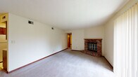 This single-floor residence offers a functional and comfortable layout designed for everyday living. The centerpiece of the main living area is a classic brick fireplace with a wood mantle, adding warmth and character to the room. The living space is generously sized, finished with neutral carpet and cream-toned walls, and features a sliding glass door that brings in natural light and provides outdoor access. A nearby dining or secondary living area connects seamlessly to the galley-style kitchen, which is well-equipped with rich wood cabinetry lining both walls, white appliances including a refrigerator and electric range, a built-in microwave hood, and light countertops over easy-care tile flooring. The kitchen also provides access to a utility area. The bedrooms are carpeted and feature neutral walls, wood-trimmed doors, and windows with blinds that allow for natural light. The full bathroom is outfitted with a single-sink vanity with wood cabinetry, a tub-shower combination, and tile flooring. Throughout the space, consistent wood baseboards and trim provide a cohesive, classic aesthetic, while textured ceilings and ample ventilation complete this well-maintained, move-in-ready home.