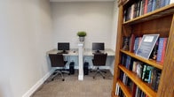 This single-floor shared workspace is thoughtfully designed to accommodate individual or collaborative computing needs in a clean, organized environment. The room centers around a built-in granite-topped desk that spans the width of the space, divided by a white decorative column that adds a subtle architectural detail while separating the two workstations. Each station is equipped with a desktop computer and monitor, paired with an ergonomic rolling chair upholstered in dark brown, ensuring comfort during extended use. A small potted green plant rests on a shelf atop the column, introducing a refreshing natural element to the otherwise neutral palette. The walls are painted in a soft gray tone, complemented by white baseboards and neutral-toned carpet underfoot, creating a calm and focused atmosphere. Natural light and recessed lighting keep the space well-lit. A warm-toned wooden bookshelf positioned at the room's entrance is stocked with a variety of books, adding both function and character. Overall, the space is efficiently laid out, balancing practicality with a welcoming, professional ambiance.