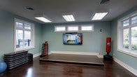 Located at 361 Darlington Avenue in Wilmington, NC, this single-floor fitness studio is thoughtfully designed to support a focused and energizing workout environment. The main exercise area features rich, dark hardwood flooring that contrasts beautifully with soft sage-green walls and crisp white baseboards and trim, creating a clean and calming aesthetic. A raised platform mat area anchors the center of the room, providing a dedicated space for guided workouts, stretching, or group training sessions. A large wall-mounted flat-screen TV is prominently positioned, ideal for streaming instructional content or fitness classes. Overhead, recessed panel lighting ensures the space is evenly and brightly lit throughout. Natural light flows in through multiple windows along the walls, including a row of clerestory windows positioned near the ceiling that add brightness without sacrificing wall space. Equipment storage is neatly integrated into the room, with a rack of weighted medicine balls and fitness mats accessible along one wall. The overall ambiance is professional yet welcoming, striking a balance between a boutique fitness studio and a functional training space suitable for individual or small group use.