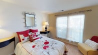 This single-floor residence offers a comfortable and inviting living environment with thoughtfully designed interiors. The bedroom is a standout feature of the space, showcasing wall-to-wall carpeting that adds warmth underfoot and a soft, cozy ambiance throughout. A large window dressed with sheer white curtains and horizontal blinds allows an abundance of natural light to fill the room, creating a bright and airy atmosphere. The neutral, warm-toned walls provide a calm backdrop that complements the room's decorative accents. A stylish wrought-iron framed mirror adorns one wall, adding an artistic and elegant touch to the space. The room is further enhanced by a compact half-moon accent table paired with a classic table lamp, offering both functionality and charm. The overall design aesthetic balances simplicity with decorative warmth, making the space feel welcoming and well-appointed. The single-floor layout ensures easy accessibility and a seamless flow throughout the home. This residence is well-suited for those seeking a cozy, low-maintenance living space with tasteful interior details.