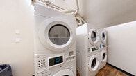 Located at 811 15th St in Greeley, Colorado, this shared laundry room is a compact, efficiently organized space designed to serve its users with practical functionality. The room features multiple stacked Whirlpool commercial-grade washer and dryer units, maximizing the available floor space by utilizing vertical stacking configurations. Each unit is coin-operated, clearly displaying pricing on digital readouts, making transactions straightforward for users. The machines are arranged along the walls, allowing for reasonable circulation through the space.

The room is finished with warm-toned luxury vinyl plank flooring that adds a clean, updated aesthetic to the utilitarian space. Neutral, light beige walls keep the interior feeling bright and tidy. A small window positioned near the ceiling allows for a degree of natural light to enter the room. Visible ventilation ducting runs overhead, supporting proper airflow for the dryer units. A self-closing, wood-panel entry door provides secure and convenient access. A waste bin is positioned near the entrance for resident convenience. Multiple light switch panels are mounted on the walls, ensuring well-distributed lighting throughout. Overall, the space is clean, functional, and well-equipped to meet shared laundry needs.