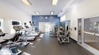 This fitness center offers a spacious, single-level layout designed to accommodate a comprehensive workout experience. The interior features a bold blue accent wall contrasting with crisp white walls and ceilings, creating a dynamic yet clean aesthetic. Durable dark rubber flooring spans the entire space, providing both safety and functionality for various exercise activities.

The cardio zone is well-stocked with multiple Star Trac treadmills, elliptical trainers, and stationary bikes, all arranged in an organized row to maximize floor space. Strength-training options are equally thorough, featuring a multi-station cable machine, a leg extension and curl station, and additional resistance equipment positioned throughout the room. A dedicated free weight area includes a full rack of dumbbells in graduated sizes, accompanied by a flat bench and a large wall-mounted mirror for form monitoring.

Natural light filters in through several windows and sliding glass doors that open to an outdoor area, enhancing the bright, energizing atmosphere. Recessed and flush-mounted fluorescent lighting ensures consistent illumination throughout. The overall layout is thoughtfully organized, offering ample room to move between equipment zones comfortably.