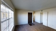 This single-floor residential space offers a straightforward and functional layout designed for comfortable everyday living. The main living area is finished with wall-to-wall carpet in a neutral tone, providing a warm and practical foundation underfoot. A textured popcorn ceiling runs throughout, adding a classic touch to the interior.

The entry is marked by a bold, dark-stained six-panel door with gold hardware, creating a strong visual contrast against the light beige walls. A small hard-surface tile or vinyl landing at the base of the entry door provides a practical transition point from the outdoors. Natural light filters in through a window along one wall, brightening the otherwise neutral interior.

Just off the main area, an open passage leads to additional spaces, while a closet with a standard rod is visible nearby, offering convenient built-in storage. The walls are painted in a clean, neutral beige throughout, keeping the space feeling open and easy to personalize. A wall vent indicates a central climate control system is in place. Overall, the space presents a clean, move-in-ready environment with practical finishes and a simple, uncluttered design aesthetic well-suited for a variety of living arrangements.