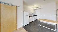 Located at 131 E Green St in the heart of downtown Ithaca, NY, this single-floor unit offers a clean, modern layout with a straightforward, functional design. The open living and dining area is generously sized, featuring warm neutral carpeting and large windows with wood-framed surrounds that frame views of the surrounding cityscape, allowing ample natural light to fill the space. Exposed ductwork along the ceiling adds a subtle industrial character to the interior.

The kitchen is efficiently arranged along one wall and includes white cabinetry, dark countertops, a full-size refrigerator, an electric range, and a sink — all designed for practical everyday use. A sliding barn-style door with natural wood finish connects the kitchen area to the rest of the unit, adding a distinctive design element.

The bedroom is carpeted and features elevated ceilings, a built-in closet with natural wood sliding doors, and direct access to the bathroom. The bathroom is finished with light ceramic tile flooring and includes a soaking tub and toilet in a clean, minimalist layout with crisp white walls throughout.

Overall, the unit presents a simple, contemporary aesthetic suited to urban living, with its downtown Ithaca location providing convenient access to local amenities, dining, and transit.