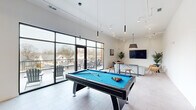 This contemporary amenity lounge offers a thoughtfully designed, open-concept layout spread across a single expansive floor. The space is defined by clean white walls, light large-format tile flooring, and elevated ceilings that lend an airy, sophisticated atmosphere throughout.

The main area serves as a versatile gathering hub anchored by a full-size pool table beneath sleek pendant lighting. Floor-to-ceiling black-framed windows and glass doors flood the interior with abundant natural light while offering direct access to an outdoor deck complete with seating and open neighborhood views.

Along one wall, a stylish bar-height counter with a built-in sink and a row of modern blue-gray stools creates a functional social and workspace zone, accented by a sage green feature wall adorned with framed botanical art and moss panels. Track lighting overhead ensures the area is well-illuminated throughout the day.

A dedicated lounge corner features a wall-mounted flat-screen TV, a plush sofa, upholstered accent chairs, and potted indoor plants that bring warmth and life to the neutral palette. Additional intimate seating nooks with minimalist chairs and side tables offer quieter spaces for conversation or relaxation. Framed artwork and lush greenery are placed throughout, tying together the modern, welcoming aesthetic of this well-appointed communal space.