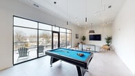 This contemporary amenity lounge offers a thoughtfully designed, open-concept layout spread across a single expansive floor. The space is defined by clean white walls, light large-format tile flooring, and elevated ceilings that lend an airy, sophisticated atmosphere throughout.

The main area serves as a versatile gathering hub anchored by a full-size pool table beneath sleek pendant lighting. Floor-to-ceiling black-framed windows and glass doors flood the interior with abundant natural light while offering direct access to an outdoor deck complete with seating and open neighborhood views.

Along one wall, a stylish bar-height counter with a built-in sink and a row of modern blue-gray stools creates a functional social and workspace zone, accented by a sage green feature wall adorned with framed botanical art and moss panels. Track lighting overhead ensures the area is well-illuminated throughout the day.

A dedicated lounge corner features a wall-mounted flat-screen TV, a plush sofa, upholstered accent chairs, and potted indoor plants that bring warmth and life to the neutral palette. Additional intimate seating nooks with minimalist chairs and side tables offer quieter spaces for conversation or relaxation. Framed artwork and lush greenery are placed throughout, tying together the modern, welcoming aesthetic of this well-appointed communal space.