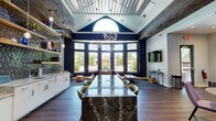 Located at 50 Lily Green Ct NW in Concord, NC, this single-floor community clubhouse delivers a sophisticated and welcoming environment defined by bold design choices and high-end finishes throughout. The centerpiece of the space is a dramatic lounge area anchored by a sweeping arc of floor-to-ceiling windows set against deep navy walls, flooding the interior with natural light and offering direct views of the outdoor pool and landscaped grounds. A sculptural, branch-inspired chandelier hangs above plush seating arrangements, creating an inviting atmosphere for relaxation and socializing. A striking granite-topped island bar runs through the heart of the space, flanked by green accent seating and open shelving with pendant lighting set against a textured metallic tile backsplash — ideal for casual gatherings. Adjacent to the bar, a dining area features a round table with upholstered purple chairs beneath exposed wood ceiling beams and a vibrant decorative wall accent. The vaulted ceiling with its distinctive corrugated metallic paneling adds architectural interest and volume to the main gathering area. Rich hardwood-style flooring flows throughout, tying each zone together cohesively. A glass-doored conference room with a sliding barn door provides a dedicated private workspace. The overall ambiance balances modern elegance with comfortable functionality.