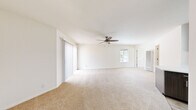 This single-floor unit offers an efficient and functional layout comprising a combined kitchen and living area, one bedroom, and one bathroom. The open living space is finished with neutral wall-to-wall carpeting and features a ceiling fan, contributing to a bright, airy atmosphere. Large vertical blinds frame a sliding glass door that opens to a private outdoor area, allowing natural light to filter through and providing outdoor access directly from the living space.

The kitchen is well-appointed with dark wood-toned cabinetry extending to the ceiling for ample storage, light countertops, a peninsula with additional counter space, stainless steel appliances including a refrigerator and dishwasher, and light wood-style flooring that contrasts neatly with the surrounding carpeted areas.

The bedroom is a comfortably sized, carpeted room featuring a ceiling fan, a sliding glass door leading to an outdoor balcony, and access to a hallway with a window for additional natural light.

The bathroom features a clean, all-white tub and shower surround with built-in corner shelving, maintaining a simple and functional design throughout. Neutral tones and consistent finishes unify the overall aesthetic of the unit.