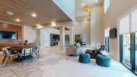This contemporary clubhouse offers an open-concept, single-story layout designed for community gathering and relaxation. The main living area impresses with soaring double-height ceilings and large sliding glass doors that flood the space with natural light while providing direct views of the surrounding outdoor landscape. Geometric hexagonal tile flooring flows throughout, adding visual interest and cohesion across each zone.

The central lounge area is furnished with modern sofas, tufted poufs in teal, and a dark wood coffee table, anchored by a woven rattan pendant light above. Adjacent to the lounge, a fully equipped kitchen bar features stainless steel appliances, dark tile backsplash, and a sleek white island with bar seating — ideal for entertaining.

A dedicated dining area is defined by warm wood-paneled walls and a wood-slat ceiling, creating an inviting, intimate atmosphere complemented by recessed lighting and decorative wall-mounted artwork. A standout alcove nook with a built-in bench seat, warm underlighting, and pendant lights provides a cozy retreat alongside a natural wood accent wall.

Throughout the clubhouse, thoughtful design details — including an ornate laser-cut wood ceiling panel, brick accent walls, and a mix of wood and tile textures — create a warm yet modern ambiance well-suited for community events and everyday use.