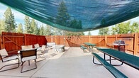 Located at 7650 Highlands View Rd in Truckee, California, this single-level outdoor space offers a well-appointed patio designed for comfortable outdoor living in the Sierra Nevada foothills. The space is fully enclosed by a rich, warm-toned redwood privacy fence, creating an intimate and sheltered environment while tall pines and mature trees visible beyond the fence provide a natural, forested backdrop beneath open blue skies.

The concrete patio is thoughtfully organized into multiple functional zones. A shade sail stretches overhead, providing relief from direct sunlight and defining a central gathering area. Multiple green picnic-style tables offer ample seating for group dining or socializing. Adjacent to this area, a gas BBQ grill is conveniently positioned along the fence line, making outdoor cooking practical and accessible.

Toward one end of the patio, a round glass-top table surrounded by cushioned chairs creates a more intimate dining nook, ideal for casual meals. A comfortable outdoor bench is positioned near a stone fire pit, which serves as a natural focal point for evening gatherings. Bicycle storage along the fence adds a practical, active-lifestyle touch. The combination of shaded seating, open-air dining, grilling facilities, and a fire pit makes this outdoor space versatile and well-suited for year-round use in the Truckee mountain environment.