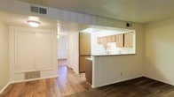 This single-story space showcases a practical and open layout that connects the main living area to the kitchen through a convenient pass-through counter design. The kitchen is equipped with light wood cabinetry offering ample storage, a white refrigerator, and a sink positioned along the counter, all visible from the adjoining living space. The pass-through breakfast bar adds functionality, making the kitchen feel connected to the rest of the unit while maintaining a defined separation between spaces.

Hardwood flooring runs throughout the main living and hallway areas, adding warmth and a sense of continuity. The hallway extending from the main space leads to additional rooms, suggesting a thoughtful floor plan that keeps living and private areas distinct. Overhead ventilation is provided through ceiling-mounted HVAC registers, and recessed and pendant lighting contribute to a well-lit interior. The textured ceiling adds a subtle design detail consistent throughout the space. Overall, the layout is straightforward and functional, offering a comfortable and efficiently organized single-floor living environment.