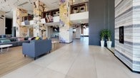 This two-floor space is defined by an open, high-energy design that blends industrial and contemporary aesthetics throughout. The main level features soaring double-height ceilings with exposed ductwork, large-format porcelain tile flooring, and sweeping floor-to-ceiling windows that frame expansive city skyline views and flood the interiors with natural light. Dramatic columns wrapped in layered mixed-media artwork serve as striking focal points, lending an artistic, urban character to the space.

The main level accommodates a variety of zones, including a spacious lounge area with modular sectional seating, a sleek blue kitchen counter with a sink, casual dining and co-working tables, and glass-enclosed meeting rooms ideal for focused collaboration. A recreation area features both a foosball and ping-pong table. Light wood paneling accents walls alongside deep navy and dark grey tones, creating a visually layered palette.

A staircase with industrial-style metal railings connects to the mezzanine level above, which overlooks the main floor through open balustrades. Throughout both levels, the design balances energetic communal areas with quieter work zones, making this a versatile and visually compelling environment.