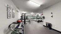 The fitness center at 21 Lakeside Ave in Pompton Lakes, NJ is a well-equipped, single-floor facility designed to support a well-rounded workout routine. The space features a clean, modern aesthetic with light gray walls, black trim accents, and durable rubber-speckled flooring throughout that provides both safety and comfort underfoot.

The cardio zone is anchored by multiple treadmills and an elliptical trainer, all positioned in front of a large wall-spanning mirror that enhances the sense of space and allows users to monitor their form. Mounted flat-screen televisions are installed above the mirror wall, adding an element of entertainment during workouts. A wall-mounted split air conditioning unit ensures climate control year-round.

The strength training area includes a multi-function cable and Smith machine with an adjustable bench, a dedicated dumbbell rack stocked with a range of free weights, and medicine balls for functional training. Motivational framed artwork decorates the walls, contributing to an energizing atmosphere.

The facility is accessed through glass-panel doors with black frames and is equipped with proper safety features including emergency exit signage and a security camera. Informational notices are posted near the entrance for user guidelines. Bright overhead fluorescent strip lighting illuminates the entire space evenly, ensuring excellent visibility throughout all workout areas.
