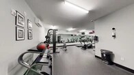 The fitness center at 21 Lakeside Ave in Pompton Lakes, NJ is a well-equipped, single-floor facility designed to support a well-rounded workout routine. The space features a clean, modern aesthetic with light gray walls, black trim accents, and durable rubber-speckled flooring throughout that provides both safety and comfort underfoot.

The cardio zone is anchored by multiple treadmills and an elliptical trainer, all positioned in front of a large wall-spanning mirror that enhances the sense of space and allows users to monitor their form. Mounted flat-screen televisions are installed above the mirror wall, adding an element of entertainment during workouts. A wall-mounted split air conditioning unit ensures climate control year-round.

The strength training area includes a multi-function cable and Smith machine with an adjustable bench, a dedicated dumbbell rack stocked with a range of free weights, and medicine balls for functional training. Motivational framed artwork decorates the walls, contributing to an energizing atmosphere.

The facility is accessed through glass-panel doors with black frames and is equipped with proper safety features including emergency exit signage and a security camera. Informational notices are posted near the entrance for user guidelines. Bright overhead fluorescent strip lighting illuminates the entire space evenly, ensuring excellent visibility throughout all workout areas.