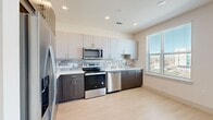 This thoughtfully designed one-bedroom, one-bathroom residence offers a clean, modern aesthetic across its single floor. The open kitchen is a standout feature, equipped with stainless steel appliances including a refrigerator, range, dishwasher, and over-the-range microwave. Two-tone cabinetry pairs dark lower cabinets with light upper cabinets, complemented by a subtle tile backsplash and light countertops. A window above the sink allows natural light to brighten the cooking space, which flows into an open living area with light wood-style flooring and a ceiling fan overhead. Sliding glass doors in the living area invite additional natural light and outdoor views. A bright, clean hallway connects the main living spaces to the private quarters, featuring recessed lighting and crisp white walls. The bedroom is carpeted for comfort, fitted with a ceiling fan, and includes a walk-in closet with built-in shelving for organized storage. A horizontal clerestory window adds a unique architectural detail to the bedroom. The bathroom offers a dark-toned vanity with ample storage, a white countertop, wall-mounted cabinet, and a tiled shower/tub combination, all set on stone-look tile flooring. The overall design is contemporary and cohesive throughout.
