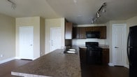 This single-floor unit offers a thoughtfully designed layout that blends functionality with a clean, modern aesthetic. The kitchen serves as a central highlight, featuring rich dark wood cabinetry that provides ample storage space both above and below the countertops. The countertops showcase a granite-style finish in warm, earthy tones, offering generous prep and serving space. A double-basin stainless steel sink is conveniently integrated into the extended kitchen counter, which doubles as a breakfast bar or casual dining surface. Black appliances, including a gas range and built-in microwave, complement the dark cabinetry for a cohesive, contemporary look. Overhead, a track lighting system provides targeted illumination throughout the kitchen area. The neutral-toned walls and light-colored ceilings create a bright, open ambiance that extends into the adjoining living areas. Two interior doors are visible from the kitchen, suggesting access to additional rooms such as bedrooms or storage spaces. The overall design balances practicality and style, making this a versatile and inviting living space suited for everyday comfort.