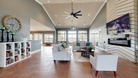 This single-floor community clubhouse offers an expansive, open-concept layout designed for gathering and relaxation. The centerpiece is a grand lounge area distinguished by soaring vaulted ceilings adorned with large-blade ceiling fans and recessed lighting, creating an airy and inviting atmosphere. Warm terracotta tile flooring runs continuously throughout, lending a cohesive, classic character to the space. The lounge features generous seating arrangements with neutral-toned sofas accented by teal throw pillows, centered around a bold floor-to-ceiling chevron-patterned fireplace surround topped with a mounted flat-screen TV. Walls of multi-pane windows and French doors bathe the interior in natural light while offering views of the outdoor surroundings. Adjacent to the lounge, a fully equipped kitchen includes stainless steel appliances, a large island with bar seating, white cabinetry, and granite countertops — ideal for events and casual gatherings. A dedicated dining area nearby features a substantial wooden table, bench seating, and a stylish pendant light fixture, with direct access to the outdoors through French doors. The overall ambiance is modern yet welcoming, with slate-blue walls, crisp white crown molding, and thoughtful décor throughout.