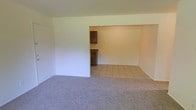 This single-floor space offers a straightforward, functional layout designed for comfortable everyday living. The living area is finished with wall-to-wall carpet in a neutral gray tone, providing a soft and inviting foundation for the room. Painted in crisp, light beige, the walls throughout contribute to a bright and airy atmosphere. An open pass-through connects the living area seamlessly to the kitchen, creating a sense of openness between the two spaces. The kitchen is finished with light ceramic tile flooring, offering a clean contrast to the carpeted living area. Dark espresso-toned cabinetry lines the kitchen walls, providing ample storage while adding a grounding visual element to the otherwise light interior. The countertops complement the cabinetry with a similarly dark finish. A door off the living area, painted white to match the trim, leads to an additional room or closet space. The overall ambiance is clean, understated, and move-in ready, with a neutral palette that offers flexibility for a variety of interior styles. The layout is efficient and well-organized, making practical use of the available floor plan on a single level.