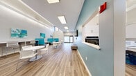 Located at 3 Village Way in Brookline, Massachusetts, this single-floor common lounge offers a thoughtfully designed, open-plan space ideal for gathering, collaboration, or relaxation. The interior is defined by a crisp white and teal color palette, with a bold teal accent wall serving as a focal point alongside a wall-mounted flat-screen television. Wood-look plank flooring runs throughout, lending warmth to the otherwise contemporary aesthetic. The layout is generously proportioned, with soaring ceilings that amplify the sense of openness. Natural light streams in through clerestory windows positioned high along the walls, as well as through large skylights and a glass exterior door that connects the space to the outdoors. Recessed and linear LED lighting supplements the natural illumination for a consistently bright environment. The seating area is versatile and well-appointed, featuring teal upholstered booth-style banquettes with integrated work surfaces, round white pedestal tables with modern white chairs, and a cushioned ottoman for flexible use. Abstract teal and blue artwork adorns the walls, complementing the overall color scheme. A built-in kitchen nook along one wall includes an oven and cabinetry, adding functional convenience. The overall ambiance is clean, modern, and welcoming, making it a highly functional and visually appealing communal environment.