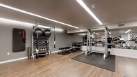 This fitness center is designed with both functionality and aesthetics in mind, offering a well-organized space for a variety of workout routines. The room features warm hardwood-style flooring that transitions to rubber matting in the free weights and strength training areas, providing both comfort and safety underfoot. Linear LED lighting runs along the smooth, neutral-toned ceiling, ensuring the entire space is brightly and evenly illuminated.

Along one wall, a sturdy metal storage rack holds an assortment of medicine balls, stability balls, and accessories, while TRX suspension straps hang nearby for bodyweight training. A central adjustable workout bench provides versatility for a range of exercises. A full dumbbell rack lines another section of the room, stocked with a complete set of weights for strength training. A power rack with pull-up bar anchors the strength training zone, flanked by large mirrors that visually expand the space and allow for proper form checks. The neutral gray walls contribute to the clean, modern atmosphere throughout. The fitness center is thoughtfully curated to support cardio warm-ups, strength training, functional fitness, and flexibility work all within a single, cohesive environment.