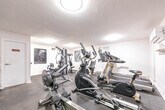 This fitness facility at 187 Park St S in Hamilton, Ontario, is a purpose-built exercise room designed to offer a complete workout experience. The space is finished with crisp white walls, dark rubber flooring, and flush-mounted ceiling lights that provide consistent, even illumination throughout. A large wall-mounted mirror spans a significant portion of one wall, visually expanding the room and allowing users to monitor their form during exercise. The facility is well-stocked with a diverse selection of equipment to support both cardiovascular and strength training routines. Cardio machines include elliptical trainers, a treadmill, a stationary bike, and a rowing machine, while strength training is served by a multi-station cable machine by Body-Solid, a flat weight bench, and a dedicated dumbbell rack stocked with a range of free weights. Medicine balls on a storage rack add further versatility. Motivational black-and-white athletic photography adorns the walls, contributing to an energetic and focused atmosphere. A split-unit air conditioner ensures climate comfort year-round. Fitness center rules are posted near the entrance, and the room is monitored by a security camera. The space is efficiently laid out to maximize usability within its footprint.