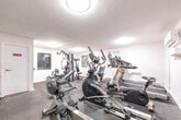 This fitness facility at 187 Park St S in Hamilton, Ontario, is a purpose-built exercise room designed to offer a complete workout experience. The space is finished with crisp white walls, dark rubber flooring, and flush-mounted ceiling lights that provide consistent, even illumination throughout. A large wall-mounted mirror spans a significant portion of one wall, visually expanding the room and allowing users to monitor their form during exercise. The facility is well-stocked with a diverse selection of equipment to support both cardiovascular and strength training routines. Cardio machines include elliptical trainers, a treadmill, a stationary bike, and a rowing machine, while strength training is served by a multi-station cable machine by Body-Solid, a flat weight bench, and a dedicated dumbbell rack stocked with a range of free weights. Medicine balls on a storage rack add further versatility. Motivational black-and-white athletic photography adorns the walls, contributing to an energetic and focused atmosphere. A split-unit air conditioner ensures climate comfort year-round. Fitness center rules are posted near the entrance, and the room is monitored by a security camera. The space is efficiently laid out to maximize usability within its footprint.