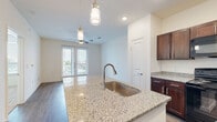 This single-floor residence offers a thoughtfully designed open-concept layout that seamlessly connects the kitchen and living areas. The kitchen is a standout feature, showcasing rich dark-wood cabinetry with sleek hardware, light granite countertops, and a suite of black appliances including a refrigerator, electric range, and built-in microwave. A central island with an undermount stainless steel sink and elegant pendant lighting serves as both a functional workspace and a visual focal point. Light wood-style flooring runs throughout the main living areas, lending warmth and continuity to the space. Double glass doors in the living area invite natural light and provide access to an outdoor area, while a ceiling fan adds comfort. The bedroom is carpeted for a cozy feel, features dual windows with blinds that frame views of surrounding greenery, and includes a ceiling fan for year-round comfort. The bathroom is finished with the same granite countertop aesthetic, rich cabinetry, a large mirror, and a tiled tub-shower combination, maintaining a consistent and polished design throughout. The overall ambiance is clean, bright, and modern, with a cohesive material palette that gives the space a refined, move-in-ready appeal.