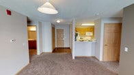 This single-floor space offers a straightforward, functional layout designed for comfortable everyday living. The living area is finished with wall-to-wall carpet in a neutral tan tone, providing a warm foundation underfoot, while a prominent pendant light fixture overhead anchors the room. A textured ceiling runs throughout, lending a consistent finish to the interior.

The kitchen is positioned to the right of the living area and is defined by a raised peninsula counter that serves as a natural divider between the two spaces. Honey-toned wood cabinetry lines the upper and lower walls, complemented by white appliances including a range and over-the-range microwave. Under-cabinet lighting adds functional illumination to the workspace.

A central hallway extends from the living area toward the front entry door, which features a classic six-panel wood design. The hallway connects to additional rooms, including what appears to be a bathroom visible to the left, finished with wood-toned cabinetry and a vanity. The overall color palette is neutral and consistent throughout, with light gray walls and white trim creating a clean, cohesive ambiance across the space.