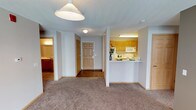 This single-floor space offers a straightforward, functional layout designed for comfortable everyday living. The living area is finished with wall-to-wall carpet in a neutral tan tone, providing a warm foundation underfoot, while a prominent pendant light fixture overhead anchors the room. A textured ceiling runs throughout, lending a consistent finish to the interior.

The kitchen is positioned to the right of the living area and is defined by a raised peninsula counter that serves as a natural divider between the two spaces. Honey-toned wood cabinetry lines the upper and lower walls, complemented by white appliances including a range and over-the-range microwave. Under-cabinet lighting adds functional illumination to the workspace.

A central hallway extends from the living area toward the front entry door, which features a classic six-panel wood design. The hallway connects to additional rooms, including what appears to be a bathroom visible to the left, finished with wood-toned cabinetry and a vanity. The overall color palette is neutral and consistent throughout, with light gray walls and white trim creating a clean, cohesive ambiance across the space.