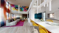 Located at 10920 Garfield Ave in South Gate, CA, this single-floor space makes an immediate impression with its dramatic double-height ceilings and an expansive, open-plan layout. The interior is defined by a striking large-scale graffiti mural—a vivid composition of oversized eyes, bold lettering, and saturated color—that commands an entire upper wall, establishing the space's energetic, artistic identity.

The main lounge area centers on a generously sized curved sectional sofa in neutral upholstery, complemented by teal and yellow accent pillows and anchored by a vibrant magenta area rug with metallic detailing. A sculptural cylindrical pendant light installation descends from the soaring ceiling overhead. Floor-to-ceiling windows dressed with colorful ombré sheer curtains allow abundant natural light while framing views of an outdoor pool and landscaped courtyard area.

Adjacent to the lounge, a sleek kitchen bar features white countertops, a teal-accented back wall, and bright yellow lattice-back bar stools. Warm wood-tone lower cabinetry and geometric tile detailing add refined texture to this zone. Light wood-look tile flooring flows throughout, unifying the entire space.

A recreation area with a yellow foosball table adds a playful element, while a pool table is visible toward the rear, reinforcing the space's social, entertainment-focused character.