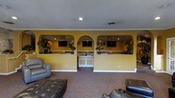 This single-floor commercial space is designed with both functionality and comfort in mind. The reception area serves as the focal point, featuring two prominent arched openings set into warm yellow walls, which frame dedicated service or check-in counters. Each counter station is equipped with mounted flat-screen monitors, making them well-suited for administrative or customer-facing operations. Decorative potted plants with vibrant red blooms are positioned throughout, adding a touch of natural color against the golden-yellow walls.

The waiting or lounge area in the foreground is furnished with dark leather seating, including plush armchairs and a tufted leather ottoman, creating a comfortable environment for visitors. The space is finished with wall-to-wall carpeting in a neutral tone, contributing to a warm and inviting ambiance. Crown molding lines the ceiling, lending a polished architectural detail to the interior. Recessed lighting provides even illumination throughout. The overall design balances a professional, organized reception layout with an approachable and welcoming atmosphere suitable for a business that regularly receives clients or guests.