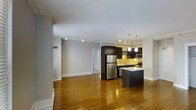 This well-appointed single-floor residence offers a thoughtfully designed open-concept layout that seamlessly connects the living and kitchen areas. Warm hardwood flooring extends throughout the main living space, adding a sense of richness and continuity to the interior.

The kitchen is a standout feature, outfitted with dark espresso cabinetry that provides a striking contrast against light countertops and a bright tile backsplash. Stainless steel appliances, including a full-size refrigerator, microwave, and range, deliver a modern, functional aesthetic. A central kitchen island with pendant lighting above serves as both a practical workspace and an informal gathering spot. Track lighting installed along the ceiling further enhances the kitchen's illuminated, contemporary feel.

The open living area is generously proportioned, offering flexible arrangement possibilities and a clean, neutral palette with light gray walls and white trim that lend the space a bright and airy ambiance. A hallway visible from the main area leads to additional rooms, suggesting a well-organized floor plan. The overall design balances modern finishes with a comfortable, livable atmosphere, making the space both visually appealing and highly functional.