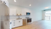 This single-floor unit showcases a clean, contemporary design throughout. The kitchen is a standout feature, outfitted with a full suite of stainless steel appliances including a refrigerator, range, built-in microwave, and dishwasher. White flat-panel cabinetry with matte black hardware lines the walls, complemented by a classic white subway tile backsplash and light-toned countertops. Light hardwood flooring runs through the kitchen and living areas, contributing to a bright, cohesive aesthetic.

The living area is open and airy, with recessed lighting overhead and a sliding glass door that leads to a balcony, allowing natural light to fill the space. An exposed metal ductwork detail adds an industrial accent that contrasts subtly with the otherwise minimalist interior.

The bedroom features plush carpeting, a large window framing outdoor rooftop and cityscape views, and a pendant light fixture. The exposed ductwork continues here, reinforcing the unit's modern-industrial character.

The bathroom is finished with large-format white floor tiles, a tiled tub-shower surround in neutral tones, a floating white vanity with an undermount sink, and a framed mirror with black trim. Matte black hardware accents are consistent throughout, tying the overall design together with a polished, uniform look.