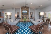 This single-floor community clubhouse showcases a thoughtfully designed open-concept interior that seamlessly blends comfort and functionality. The space is anchored by a dramatic floor-to-ceiling stacked stone fireplace, which serves as the centerpiece of the main lounge area. Vaulted ceilings with exposed beams and recessed lighting add architectural depth and a sense of spaciousness throughout.

Rich, dark hardwood flooring runs throughout the entire space, providing a warm contrast to the light gray walls and a bold terracotta accent wall near the reception area. The lounge zone features contemporary gray sofas, tufted leather accent chairs, and a striking geometric blue area rug, creating an inviting and modern gathering space.

Adjacent to the lounge, a dedicated dining area offers a round table with upholstered seating beneath vibrant pendant lighting, positioned alongside multiple sets of French doors that flood the space with natural light and provide direct access to the outdoors. A built-in workstation area with desk space and computers offers a practical business center component.

A glimpse into an adjoining fitness room reveals exercise equipment, adding to the multi-functional nature of this amenity space. The overall ambiance is modern, welcoming, and well-appointed, making it an ideal communal hub.