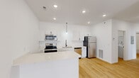 This single-floor unit presents a clean, contemporary design characterized by an all-white palette, light hardwood-style flooring, and ceiling fans throughout. The kitchen serves as a central highlight, featuring crisp white shaker cabinetry, white quartz countertops, and a full suite of stainless steel appliances, including a refrigerator, electric range, over-the-range microwave, and dishwasher. Recessed lighting and a pendant light above the sink provide layered illumination, while an extended kitchen island offers additional prep and dining space. The layout transitions smoothly into the living areas, where multiple rooms benefit from natural light through standard windows and ceiling fans for year-round comfort. A standout architectural detail is the sliding barn door, adding a rustic-modern touch to one of the bedroom or closet entryways. Bedrooms are well-proportioned with clean white walls and hardwood-style flooring. The bathroom includes a full soaking tub with shower combination. A dedicated laundry area houses a full-size top-loading washer and dryer with overhead wire shelving for storage. The overall ambiance is bright, clean, and move-in ready.
